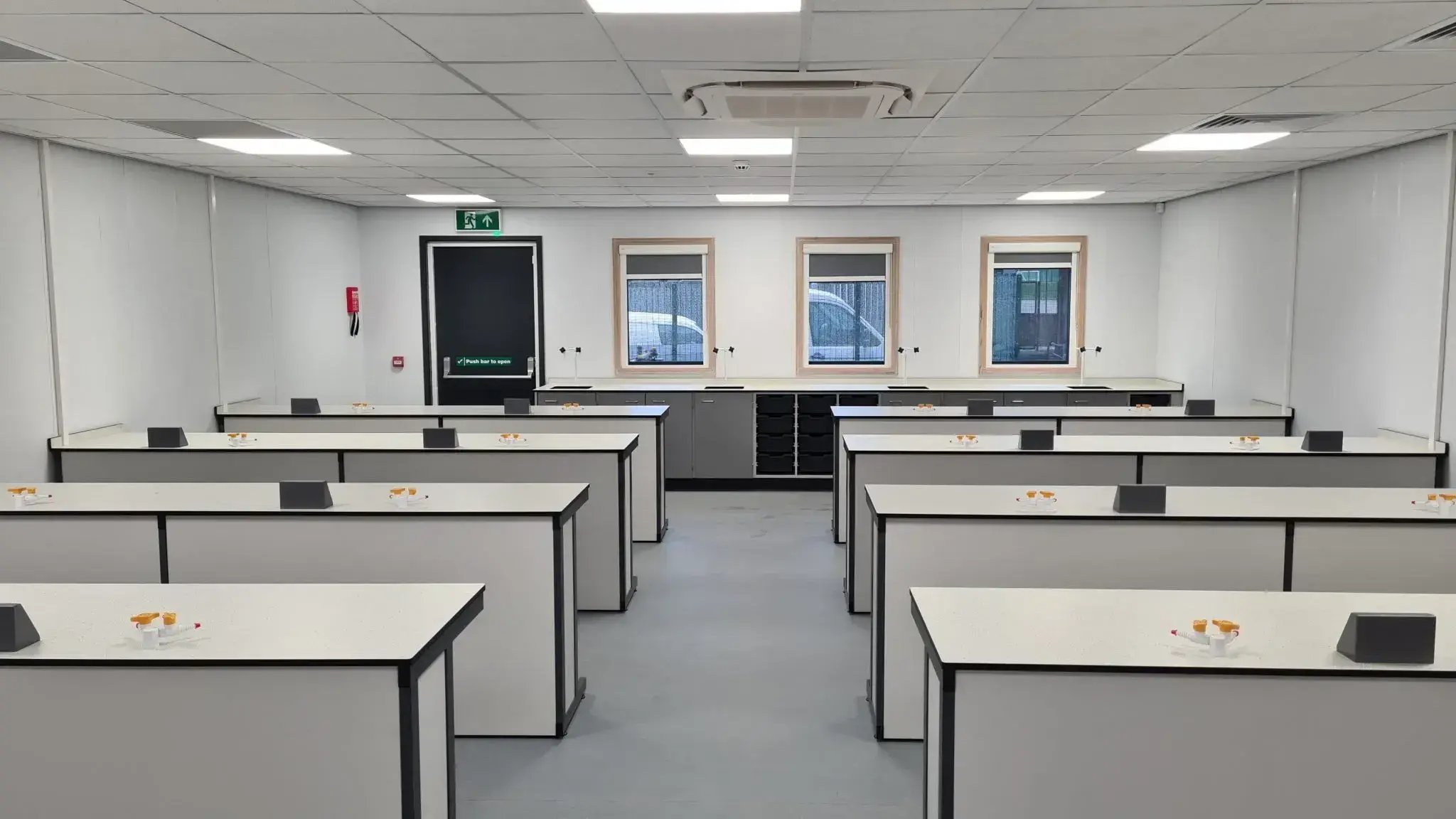 Spacious modern science laboratory classroom with rows of white lab benches, bright overhead lighting, integrated workstations, and large windows designed for practical science lessons and student experiments.