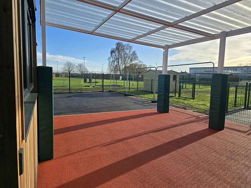 Covered outdoor play and learning area attached to a modular SEN building, featuring red rubber safety flooring, steel canopy, padded support posts,
