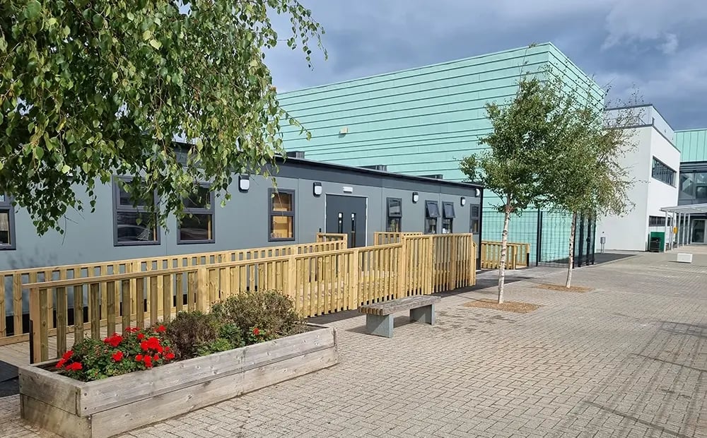 Modular classroom building at Ysgol Gogarth School, featuring accessible wooden ramp, landscaped planters, and paved outdoor learning area alongside the main school building.