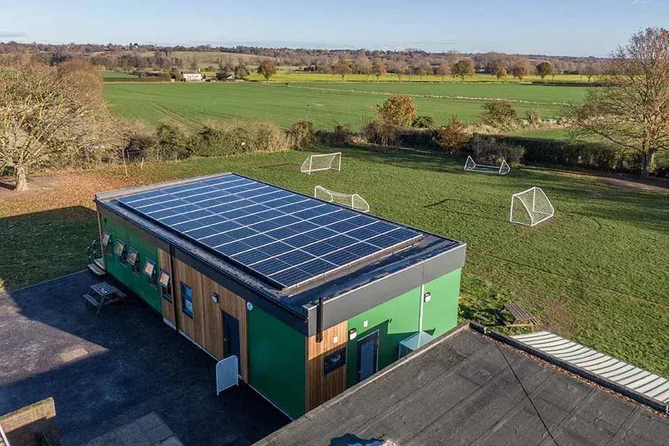 Modern sustainable modular school building with rooftop solar panels overlooking sports fields, designed for energy efficiency, renewable power generation, and environmentally friendly educational spaces.