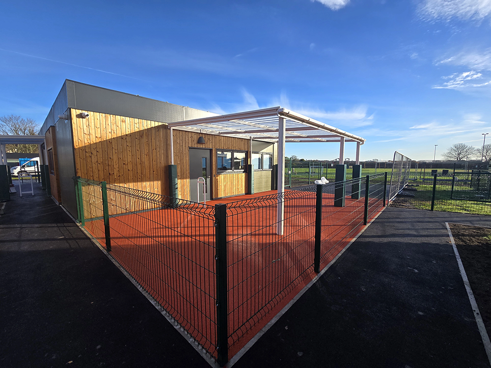 Prefabricated modular school building with wooden exterior, steel canopy, and secure fenced outdoor learning space.