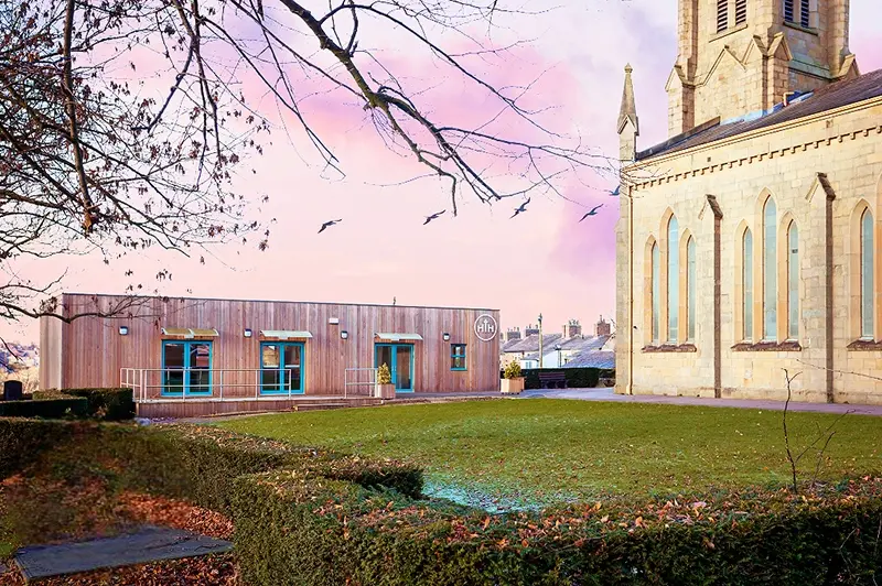 Exterior of Hurdsfield Community Hub, a modern single-storey modular community building with wooden cladding and teal-framed windows.