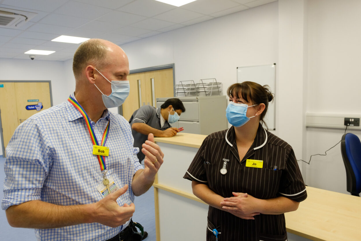 Healthcare staff wearing face masks talk together at a reception desk inside a modular building clinical setting, illustrating a calm, well-organised healthcare environment.