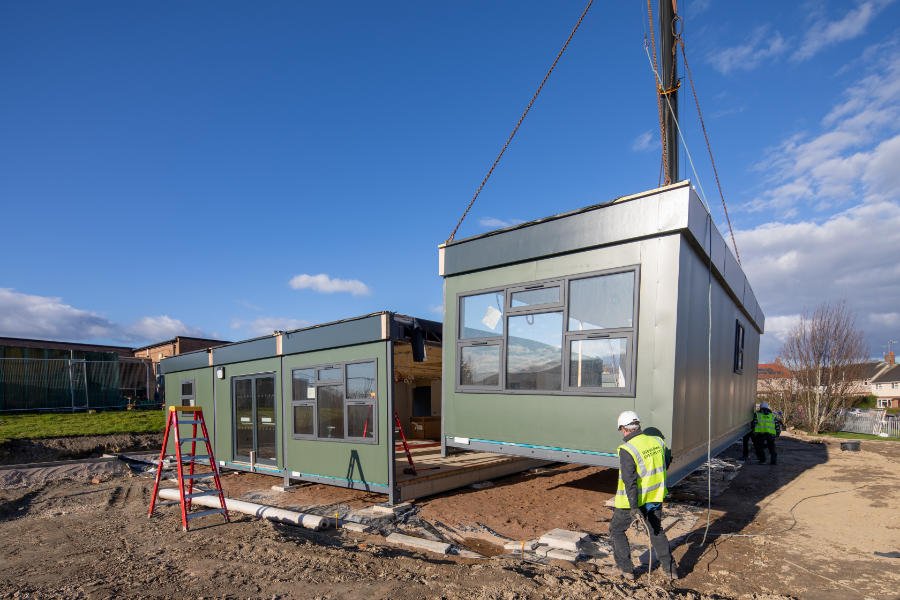 Two green modular buildings shown being put into place on site. One is already on the ground, the other is beig craned into position guided by a man in high vis gear ad white hard hat