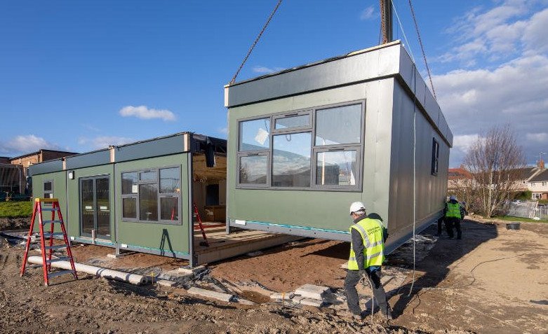 Two dark green modular buildings installed, one already secured to the ground and the other is being craned into position, guiding by workmen in high vis gear and white hard hats. This is part of our circular economy.