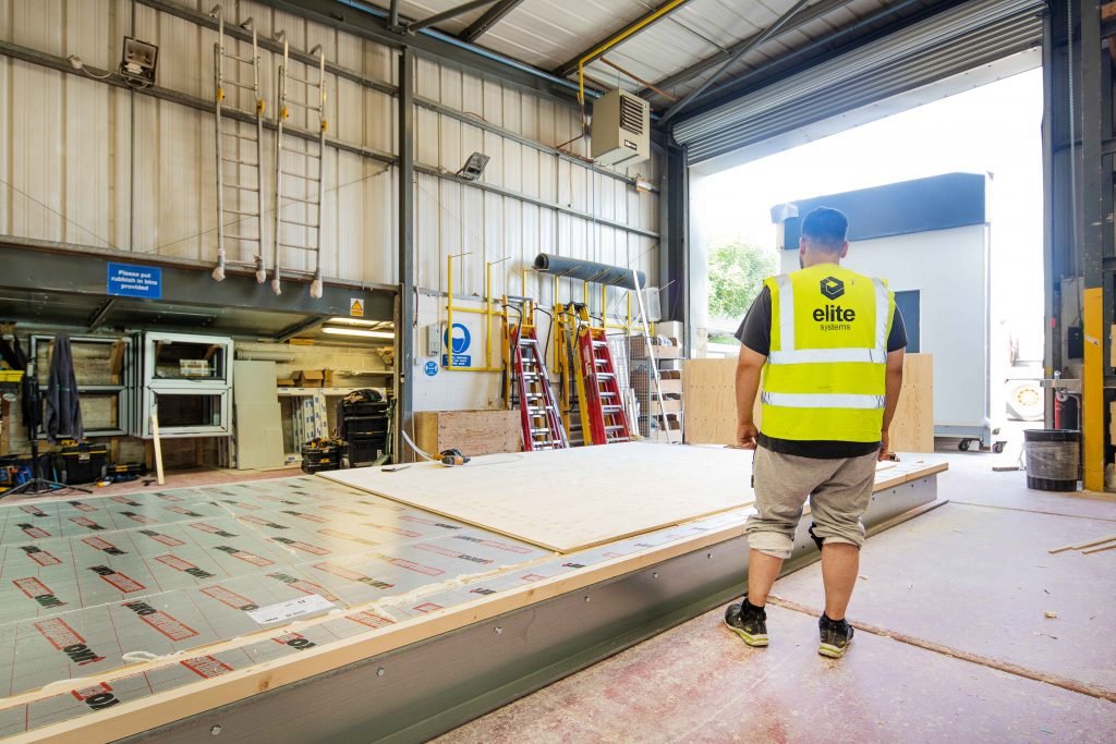 Image shows a factory floor with a flat-pack modular building whilst another is leaving the factory. A factory worker stands in high vis clothing watching