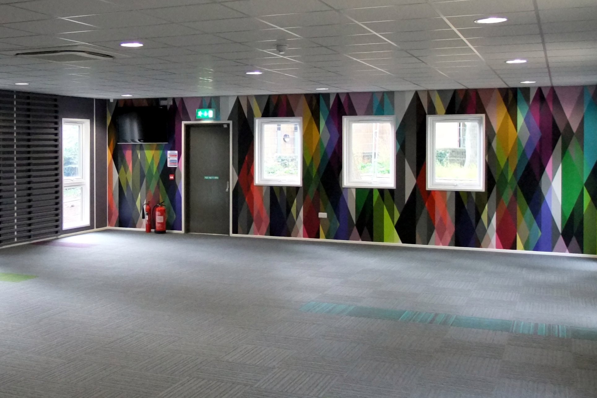 Image shows a large empty room with a multi-coloured feature wall which is made up of gemoetric shapes. The ceiling is made of ceiling tiles and there's a grey carpet covering the floor. There's 3 waist-height windows on the far wall and a floor-to-ceiling window on the far left shocasing the amount of light coming in. This was a modular built room made by Elite Systems for Sheffield Hallam University