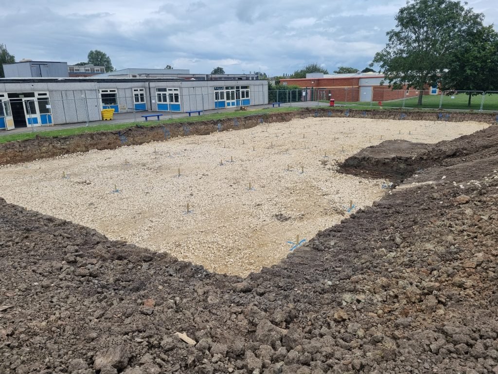 Here shows a gravel-looking bed with wooden markers evenly spaced to calculate where the piling for the foundations will go.