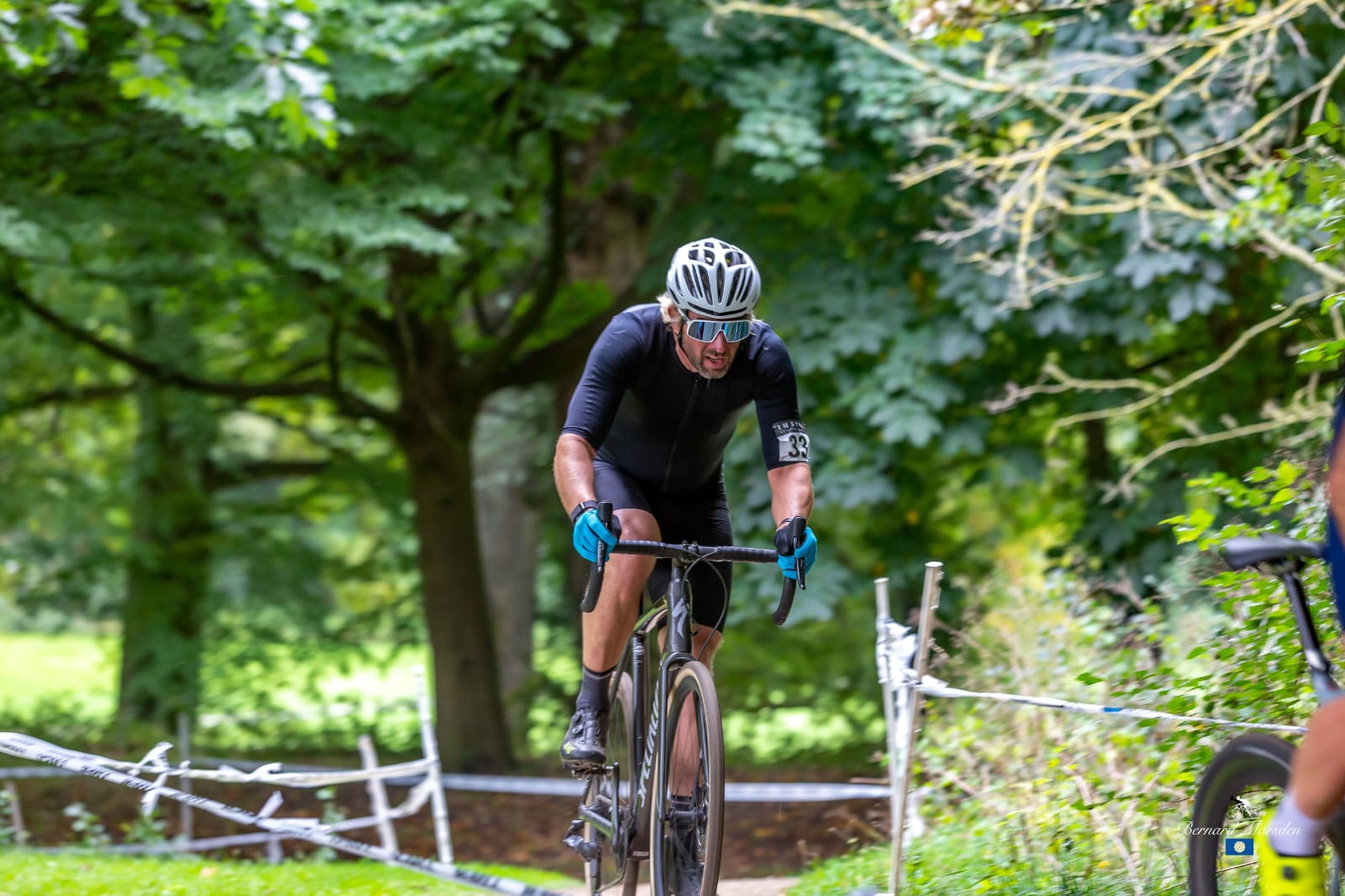 Photo shows Marcus Sutcliffe cycling cross country on a road, with trees behind.