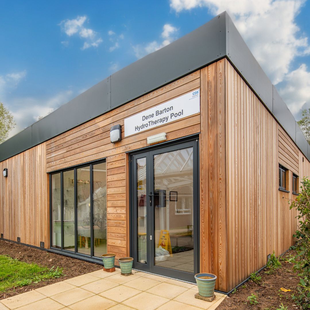 Picture shows a larch cladded building with dark grey windows and doors with a view inside to the reception space. The sign above the door says Dene Barton Hydrotherapy pool.